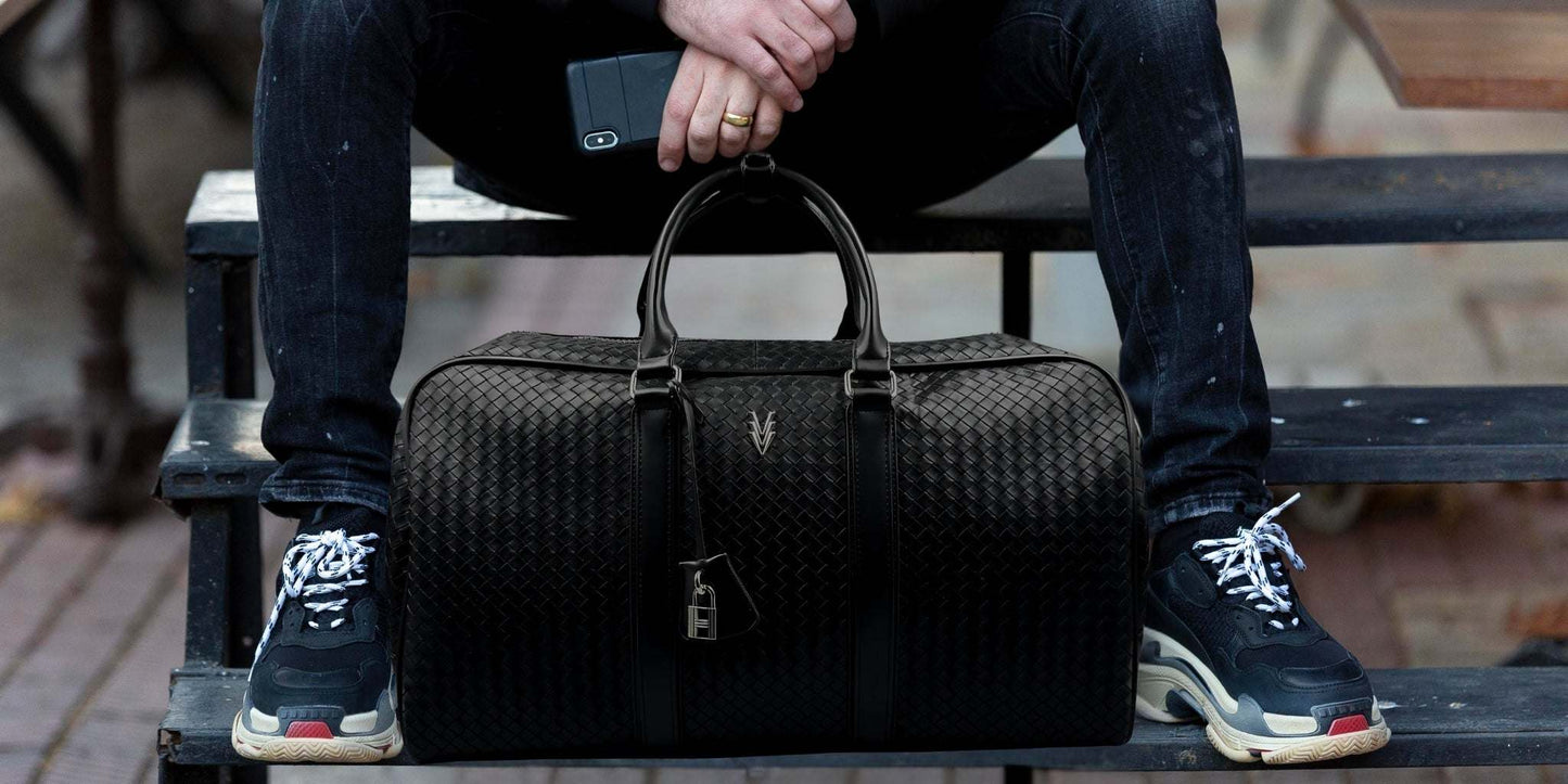 Black Rocco Weekend Duffle bag with embossed woven texture in vegan leather, held by a person sitting on outdoor steps.