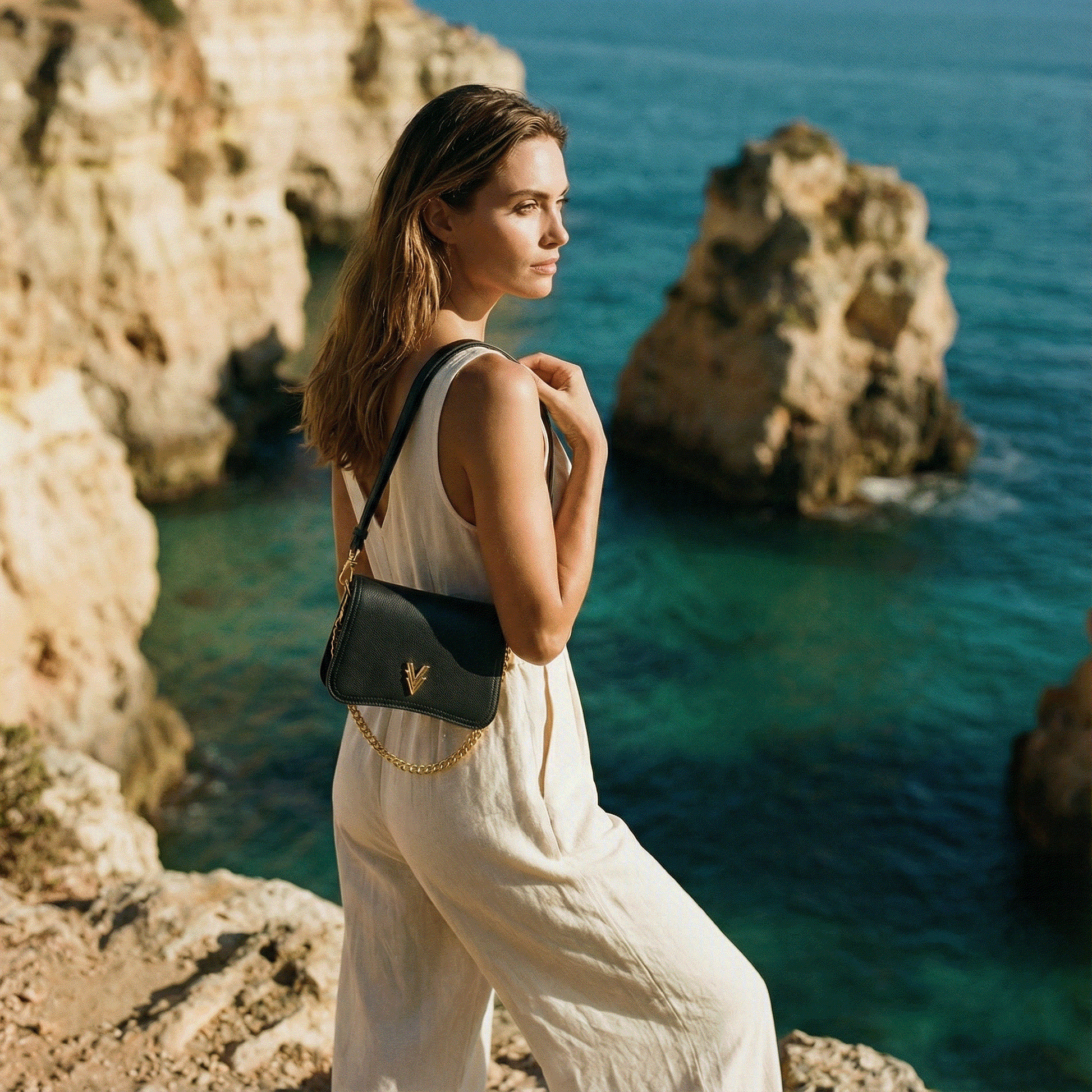 Woman in a white dress with a black handbag standing on a rocky cliff overlooking the ocean.