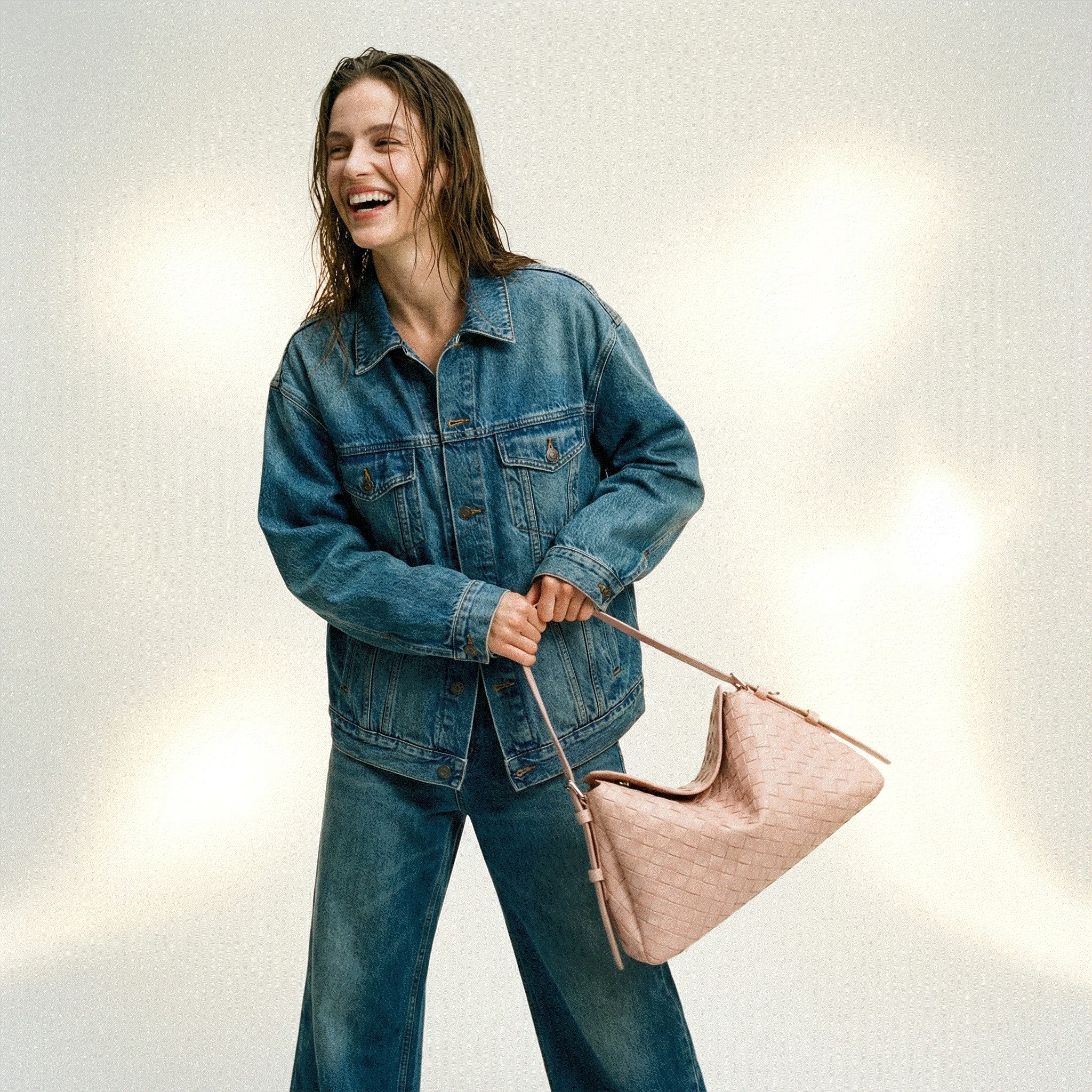 Woman wearing a denim jacket and jeans holding a pink handbag against a neutral background