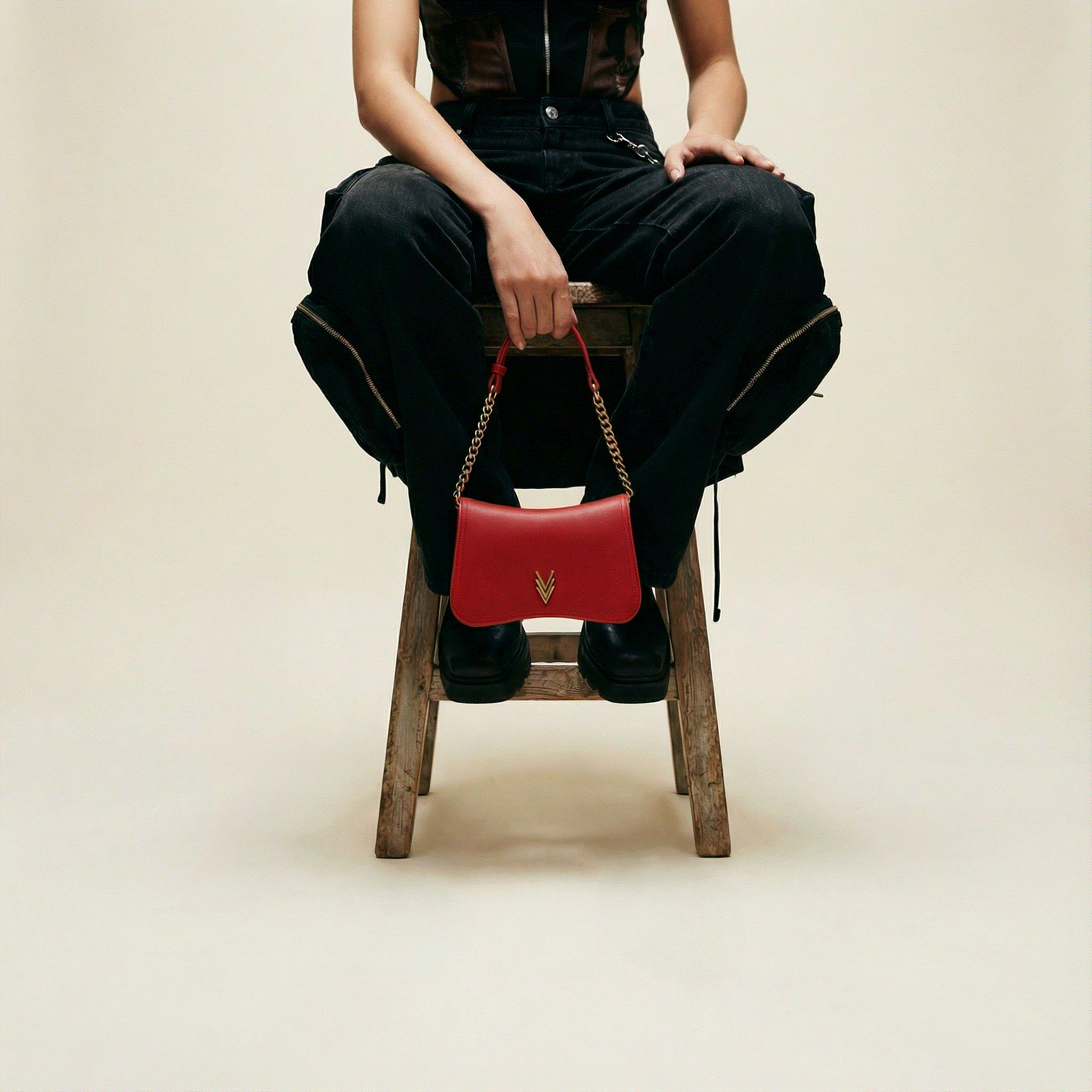 Person sitting on a wooden stool holding a red handbag against a beige background